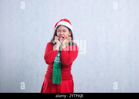 gesture of young Asian ladies wearing Christmas sweater, Santa hat, and scarf, screaming looking at camera, both hands holding cheeks, isolated on whi Stock Photo