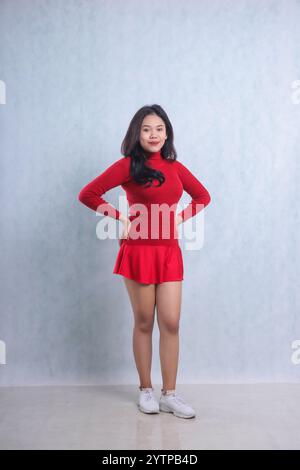 full body gesture of young Asian woman wearing red sweater, standing smiling to camera both hands holding waist, isolated on white background Stock Photo