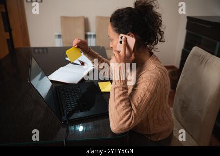 A woman sits at her dining table, balancing her phone and laptop while working from home. She is actively taking notes and managing multiple tasks sim Stock Photo