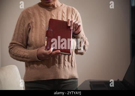 Close-up of a person holding a leather planner, ready to organize tasks beside a laptop. Perfect for illustrating productivity, planning, and office s Stock Photo