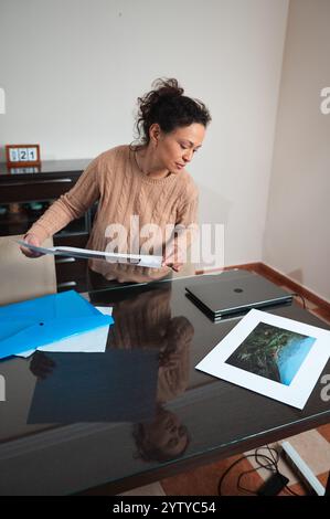 A woman in a cozy sweater reviews printed photographs at her desk, organizing with folders beside a laptop. The workspace is tidy and minimalistic, su Stock Photo