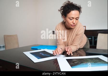 A woman closely examines printed photographs on a table, suggesting focus and concentration. The scene includes a laptop, indicating a home or office Stock Photo