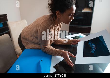 A woman examines photography prints on a desk, showcasing creativity and focus in a home office. The image captures a moment of artistic involvement a Stock Photo
