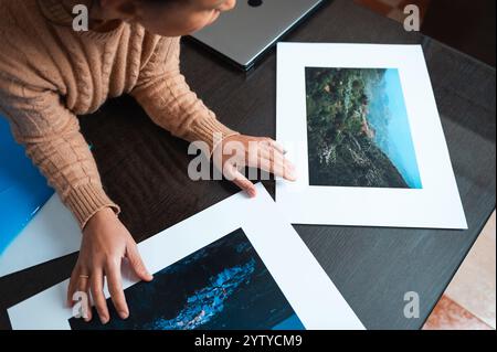 A woman examines printed nature photos on a dark wooden table, conveying creativity and focus. Stock Photo