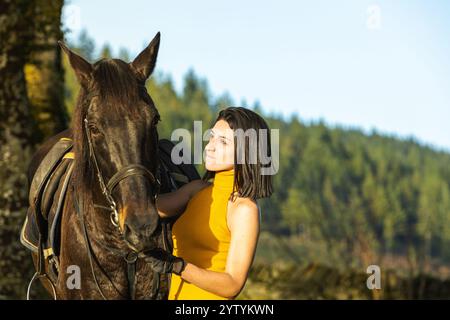 A woman in a mustard top and gloves gazes at her black horse in a serene countryside backdrop. The image captures their connection amidst lush greener Stock Photo