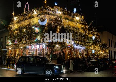 London, UK. 7th December, 2024. A black cab passes in front of The Churchill Arms pub decorated with Christmas lights. The Churchill Arms has been dec Stock Photo