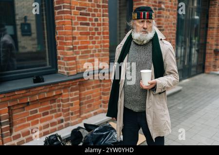 Portrait of senior adult homeless man with lengthy white beard and poor attire, clutching white paper cup while standing by his possessions on city Stock Photo