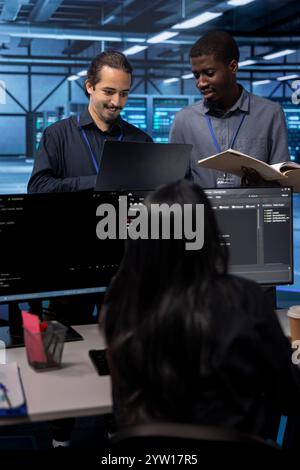 Cheerful teamworking engineers in data center looking for firewall misconfigurations affecting servers. Happy multiethnic employees checking systems bottlenecks leading to sluggish data transfer rates Stock Photo
