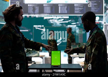Diverse team of soldiers examine information on a green screen, working with a tablet in military base of operations. Skilled IT engineers monitoring activity in control center, high tech. Stock Photo