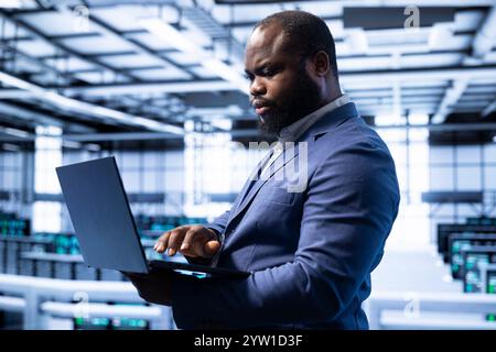 Engineer in server room establishment installing newly ordered storage units using laptop. African american man working in data center facility housing storage rackmounts Stock Photo