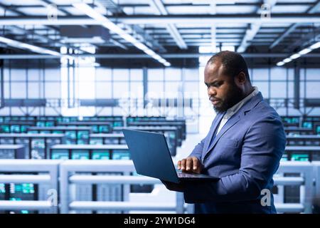 Worker in data center using laptop to configure network infrastructure such as routers, switches and firewalls. Data researcher working in server room, looking for technical malfunctions Stock Photo