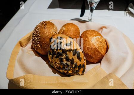 A basket of assorted bread rolls, including sesame seed, plain, and pumpkin seed varieties, served on a cloth napkin in a warm dining setting. Stock Photo