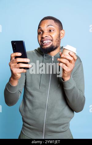Confident optimistic man browsing his smartphone and serving coffee, feeling happy after receiving good news and socializing with his friends via text messages. Modern guy with a refreshment. Stock Photo