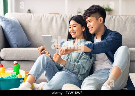 Loving asian couple with cleaning tools resting, using digital tablet Stock Photo