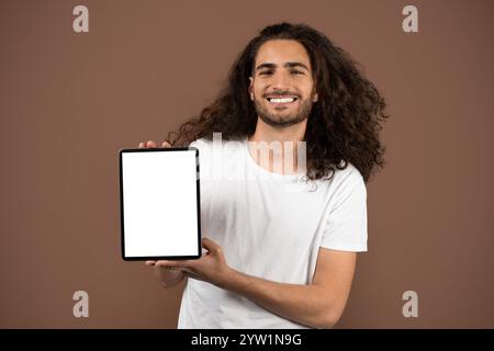 Young man with curly hair joyfully presenting tablet against neutral backdrop Stock Photo