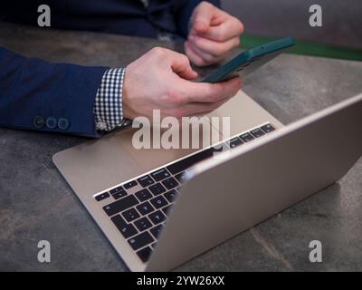 Focused businessman in modern office environment, using smartphone and laptop while dressed in blue suit and checkered shirt Stock Photo