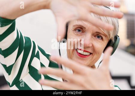 Joyful Senior Woman Making Frame with her Hands While Listening to Music with Headphones, Smiling and Looking at Camera. Stock Photo