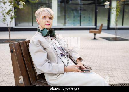 Elegant Senior Woman with Short Hair Wearing Headphones and Using Smartphone While Sitting on Bench Outdoors in City, Active lifestyle for Older adult Stock Photo