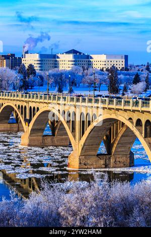 A bridge over a river with snow on the trees and a building in the background. The bridge is covered in ice and snow Stock Photo