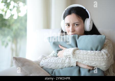 Sad asian woman wearing headphone listening audio looking hugging pillow at you at home Stock Photo