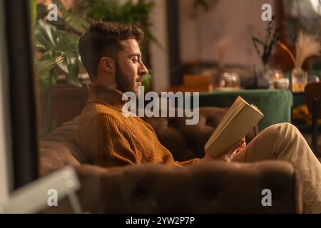 Young man in turtleneck sweater reading book at cozy cafe, side profile through window Stock Photo