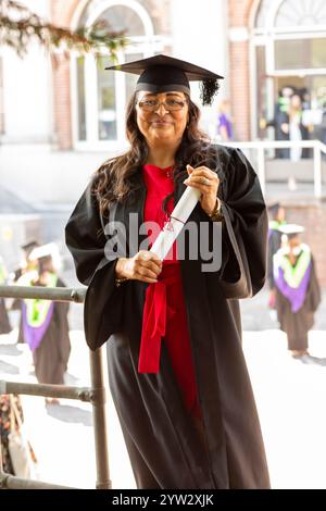 A proud graduate in cap and gown holds a diploma with a red ribbon, smiling on her graduation day with others in the background, Bournemouth, Dorset UK Stock Photo