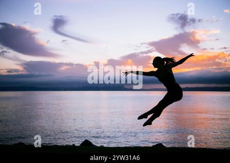 Silhouette of a woman leaping joyfully at sunset with a serene water backdrop and a vibrant sky. Stock Photo