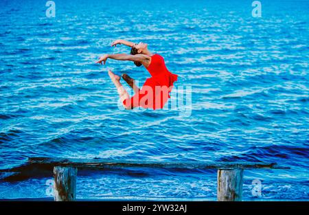 Graceful ballet dancer in a red dress performing a jump over blue water with a wooden structure in the foreground, USA Stock Photo
