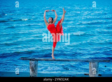 A dancer in a striking red dress performs a ballet pose on a wooden jetty against a deep blue ocean backdrop, USA Stock Photo