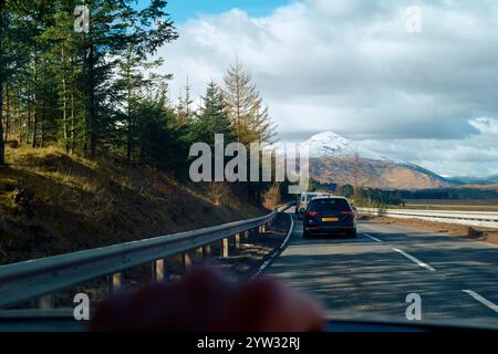 Car driving on a road with a snow-capped mountain in the background and pine trees lining the route under a blue sky with clouds, Highlands, Scotland Stock Photo
