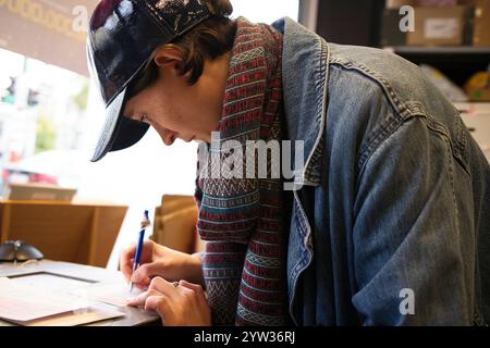 A young adult woman in a denim jacket and a baseball cap writes on a piece of paper at a table, concentrating intently. Stock Photo