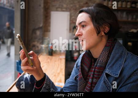 Woman sitting by a window on a wooden counter, intensely focusing on a smartphone in hand, dressed in a denim jacket and patterned sweater. Stock Photo