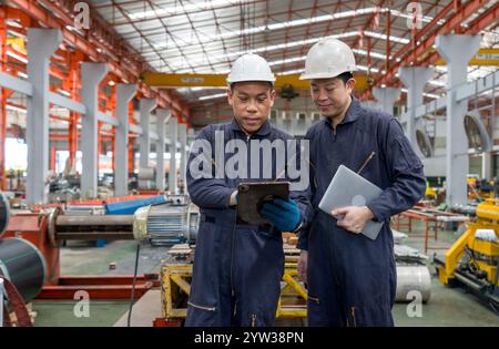 Two workers in safety helmet and mechanic coverall dust protection suit look at a tablet in a factory. They check information and discuss their task. Stock Photo