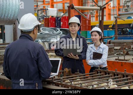 Three workers in a factory discuss a project using laptop computer. Everyone wearing protective hardhat. Atmosphere in manufactory with large machiner Stock Photo