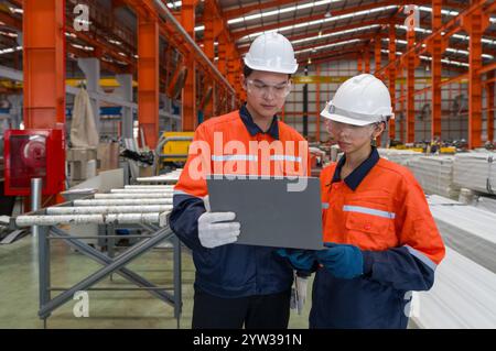 Two worker in safety gear look at a laptop computer, standing in a large factory. They are checking equipment and discussing plan. Stock Photo