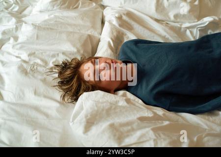 Young adult lying in bed with messy hair and a pensive expression, on a white duvet, Norway Stock Photo