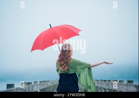 Red-haired woman holding a red umbrella on a foggy wooden pier, extending her arm out as if testing the weather. Stock Photo