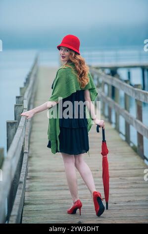 Stylish woman with a red hat and shoes holds an umbrella while standing on a wooden dock by the water. Stock Photo