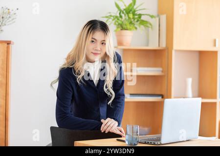 Professional young woman standing behind a desk with a laptop and a glass of water, looking at the camera with a slight smile. Stock Photo
