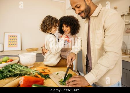 A joyful family enjoys quality time together in the kitchen, preparing a healthy meal. The boy is sitting on the kitchen table playing with a red toy Stock Photo