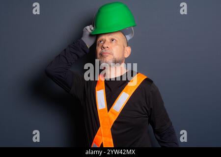 Photo of a civil worker in gloves and reflective vest putting on holding green helmet on his head. Isolated on gray background. Construction worker. Stock Photo