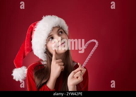 Smiling girl hold Christmas candy cane and wear red Santa Claus hat as Snow White portrait in red background Stock Photo
