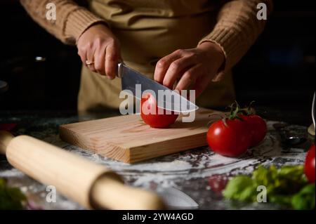 A person skillfully slicing fresh tomatoes on a wooden board in a home kitchen. The scene highlights culinary skills, home cooking, and the use of fre Stock Photo