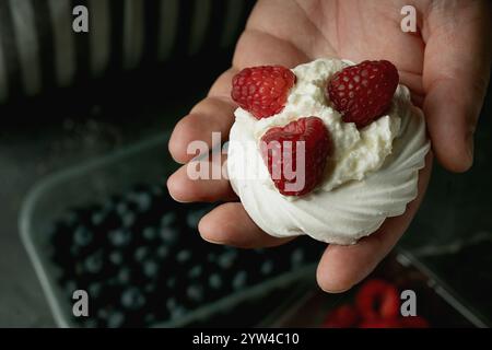 Hand assembling a meringue dessert with raspberries and blueberries, placed over whipped cream on a dark surface. Fresh berries in transparent boxes a Stock Photo