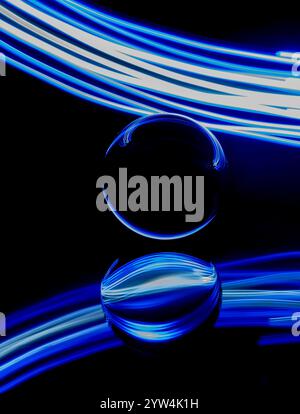 Abstract blue and white light trails created by light painting around a static glass ball set on a mirror against a plain black background Stock Photo