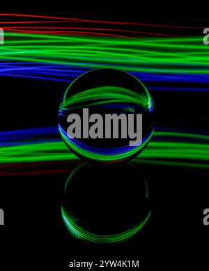 Abstract blue, green and red light trails created by light painting around a static glass ball set on a mirror against a plain black background Stock Photo