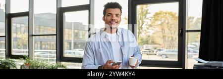 Handsome man holds a cup and smartphone, smiling in a lively, sunlit cafe. Stock Photo
