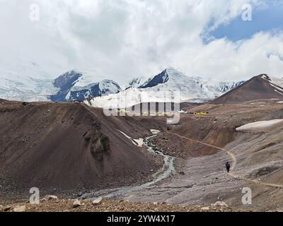 A hiker walks along a winding trail near a flowing stream with tents visible amidst majestic mountains. Stock Photo