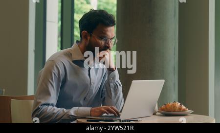 Arabian Indian man typing laptop keyboard concentrated business guy male businessman at cafe table online work computer browsing internet write tasks Stock Photo