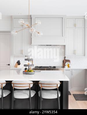 A kitchen with light grey cabinets, gold fixtures, white mosaic tile backsplash, and bar stools sitting at a large grey island. Stock Photo